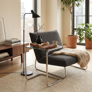 May include: A charcoal gray upholstered lounge chair with a chrome tubular frame, featuring a glass-topped side table attached to the frame. A beige faux fur throw rests on the seat, and a black vase with dried branches sits on the glass top.