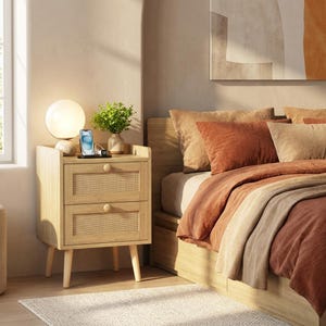 May include: A light wood nightstand with two drawers featuring woven cane panels and round wooden knobs. The nightstand stands on four tapered legs and holds a spherical white lamp, a small potted green plant, and a smartphone on a charger.
