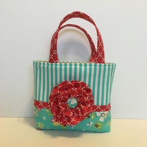 May include: A small turquoise and white striped tote bag with a red fabric flower and a white button in the centre. The bag has a red and white patterned handle.