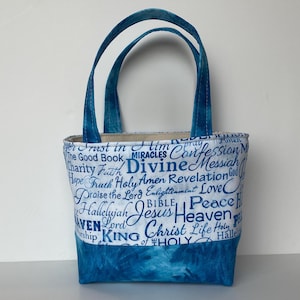 May include: A small tote bag with blue handles and a blue bottom. The bag's white fabric is covered in blue script with words like "Divine", "Jesus", and "Heaven". The bag is designed to be carried by hand.