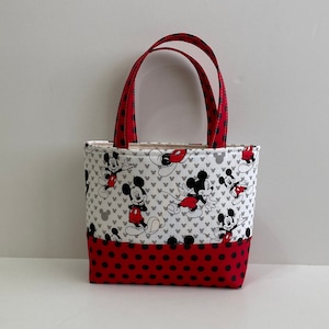 May include: A small red and white tote bag with a Mickey Mouse print. The bag has red handles and a red bottom with white polka dots.