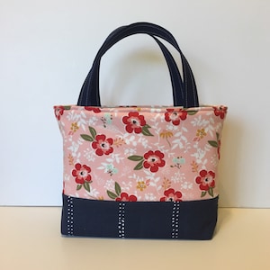 May include: A small tote bag with a floral pattern in pink, red, and white. The bottom section is navy blue with vertical white dotted lines. It has navy blue handles.