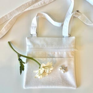 May include: A white crossbody bag with a zippered pocket and a long, adjustable strap. A white flower and a pearl brooch adorn the front. The bag is made of a textured fabric.