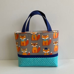 Könnte beinhalten: Eine kleine graue Tragetasche mit einem blauen Griff. Die Tasche ist aus Stoff und hat ein Muster aus Füchsen, die Brillen, Krawatten und Hüte tragen. Der Boden der Tasche ist aus türkisfarbenem Stoff mit weißen Punkten.
