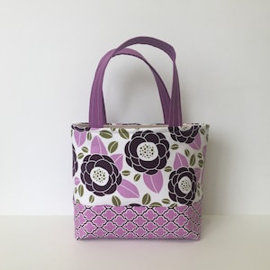 May include: A small tote bag with purple handles. The bag features a floral pattern with large dark purple flowers, green leaves, and light purple leaves on a white background. The bottom section has a repeating light purple geometric design.