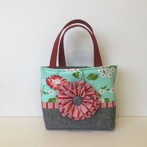 May include: A small, teal blue tote bag with a red and white striped flower embellishment. The bag has red handles and a grey fabric bottom.