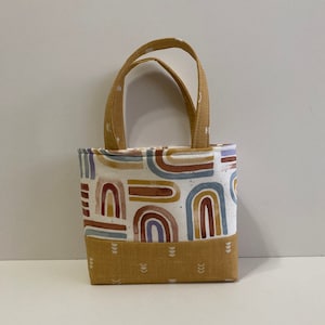 May include: A small, mustard yellow tote bag with a white fabric lining. The bag has a geometric pattern of colorful rainbows on the front. The bag has two handles.