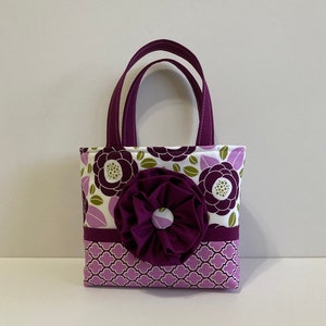 May include: A small purple and white floral tote bag with a large purple fabric flower on the front.