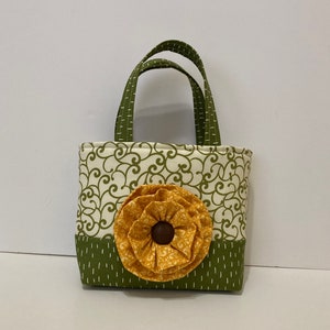 May include: A small green and white fabric tote bag with a yellow fabric flower on the front. The bag has a green fabric handle.