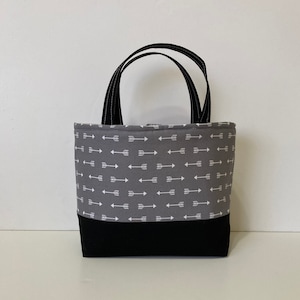 May include: A gray tote bag with black handles and a white arrow pattern. The bottom of the bag is black.