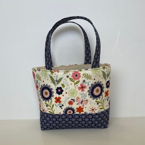 Puede incluir: Una pequeña bolsa de mano blanca con estampado floral y ribete azul marino. La bolsa tiene dos asas azul marino.
