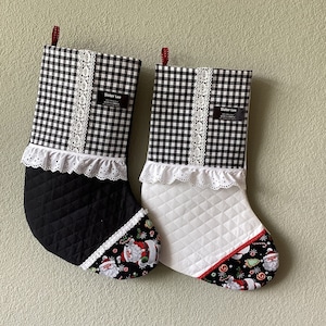May include: Two black and white gingham Christmas stockings with white lace trim. One stocking is black with a Santa Claus print on the toe and the other is white with a Santa Claus print on the toe. Both stockings have a red ribbon loop at the top and a label that says "Sister Lee".