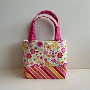 Könnte beinhalten: Eine kleine rosa Tragetasche mit weißen Punkten an den Henkeln. Die Tasche ist aus Stoff mit einem Blumendruck mit rosa, gelben und grünen Blumen, Bienen und Schmetterlingen. Der Boden der Tasche ist aus gestreiftem Stoff mit rosa, roten, gelben und orangen Streifen.