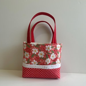 May include: A small red tote bag with white daisy floral print and a red and white checkered bottom. The bag has two red handles.