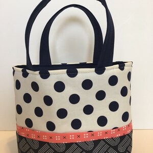 May include: A small white tote bag with black polka dots and a pink ribbon. The bottom of the bag is black with a white pattern.