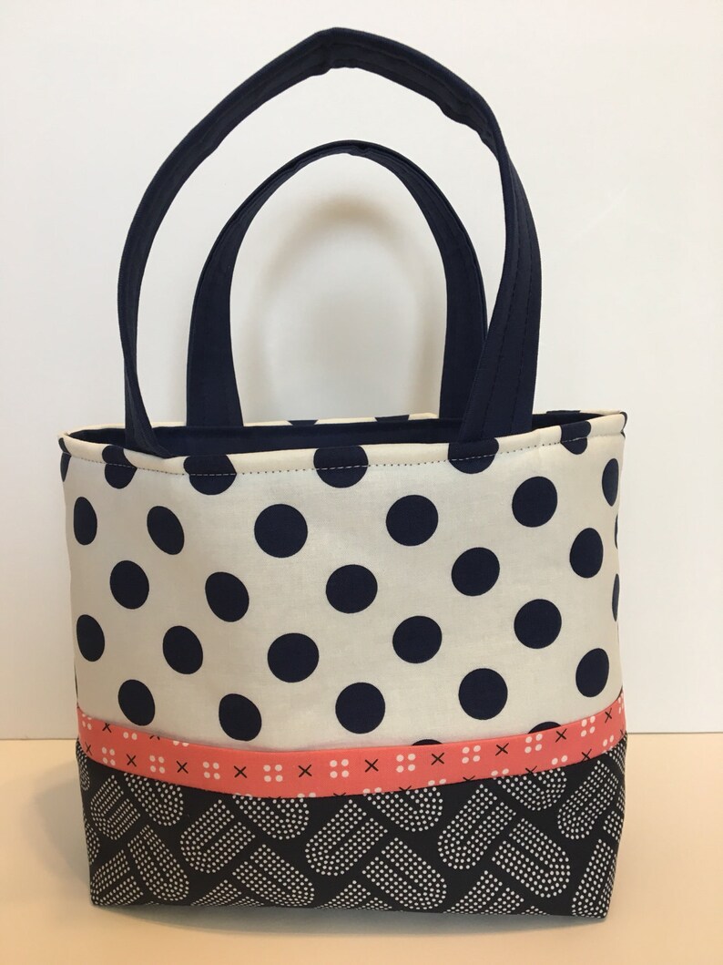 May include: A small white tote bag with black polka dots and a pink ribbon. The bottom of the bag is black with a white pattern.
