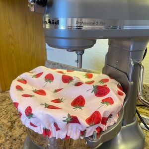 May include: A silver KitchenAid Professional 5 Plus stand mixer with a white and red strawberry print bowl cover. The cover has a ruffled edge and is patterned with red strawberries and green leaves on a white background.