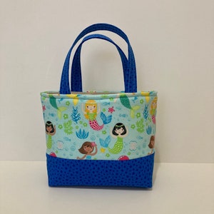 May include: A small tote bag with blue handles and a blue base. The main body of the bag is light blue and features a pattern of mermaids, fish, and aquatic plants. The handles are a solid blue with a dotted pattern.