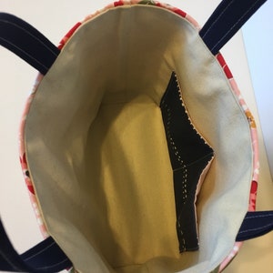 May include: A floral tote bag with a light beige interior and a black pocket. The exterior features a pink and red floral pattern with navy blue handles. The bag is designed for carrying items.