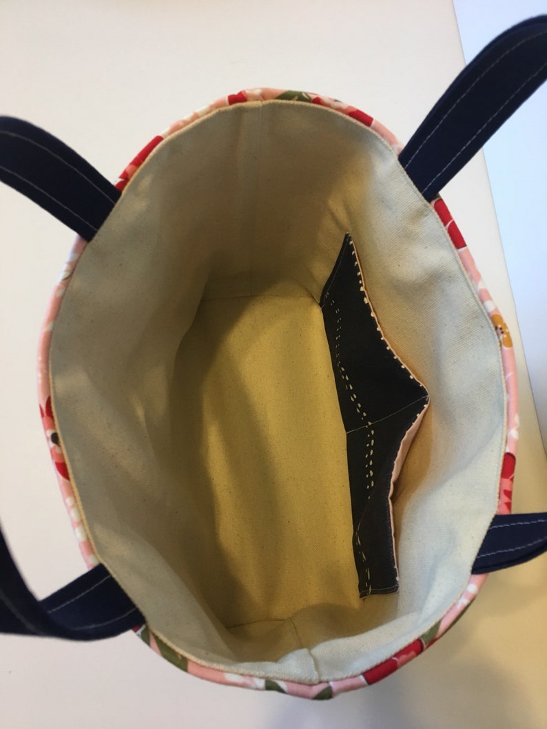 May include: A floral tote bag with a light beige interior and a black pocket. The exterior features a pink and red floral pattern with navy blue handles. The bag is designed for carrying items.
