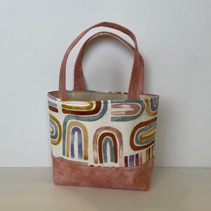 May include: A small pink tote bag with a white fabric lining and a colorful abstract rainbow pattern. The bag has two pink handles.