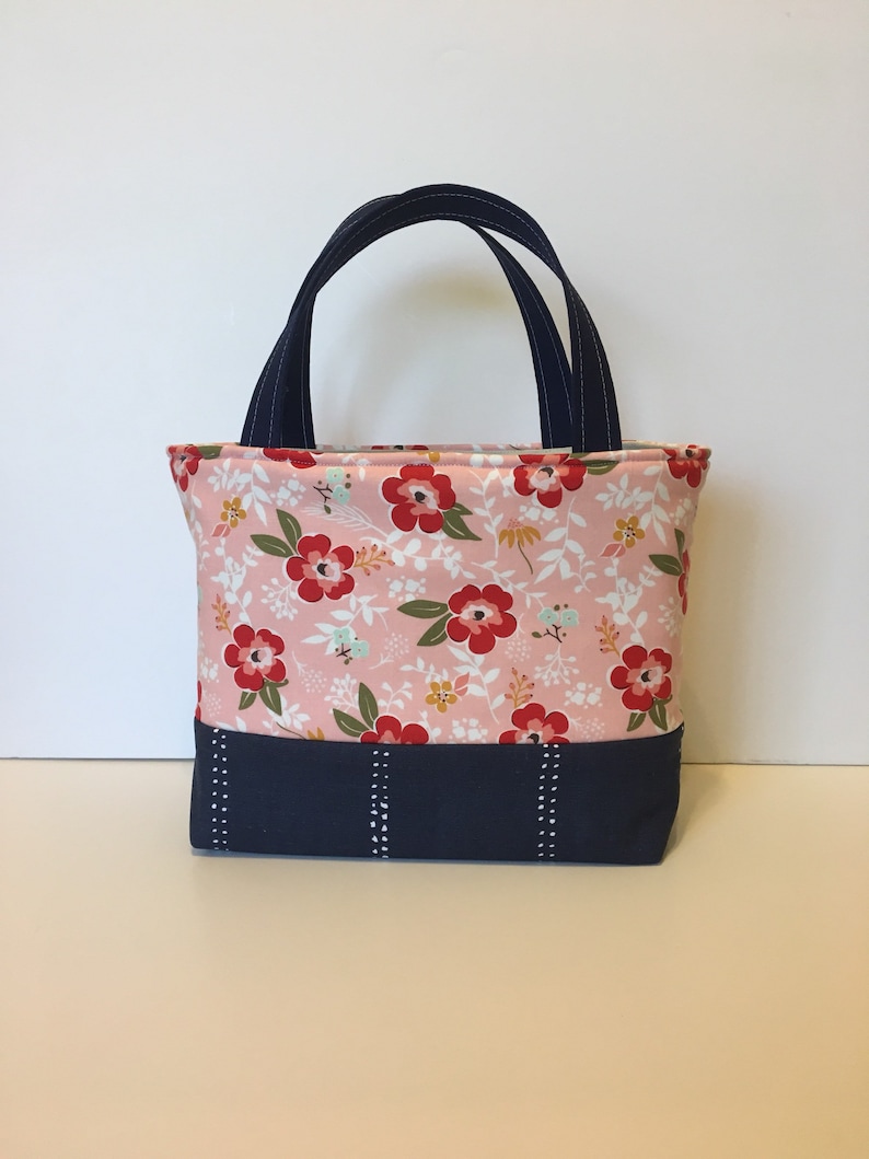 May include: A small tote bag with a floral pattern in pink, red, and white. The bottom section is navy blue with vertical white dotted lines. It has navy blue handles.