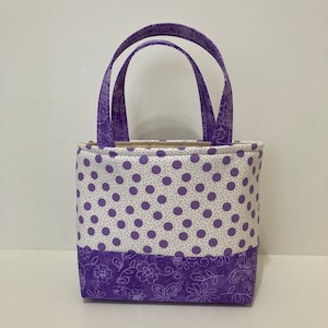 May include: A small purple tote bag with white polka dots and a floral pattern. The bag has two purple handles.