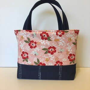 May include: A small tote bag with a floral pattern in pink, red, and white. The bag has navy blue handles and a navy blue base with white stitching details. The bag is designed for carrying small items.