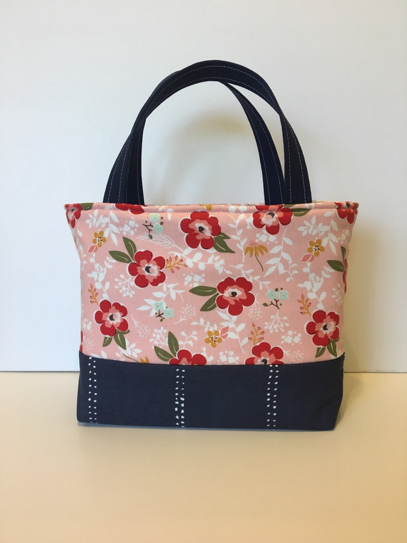 May include: A small tote bag with a floral pattern in pink, red, and white. The bag has navy blue handles and a navy blue base with white stitching details. The bag is designed for carrying small items.
