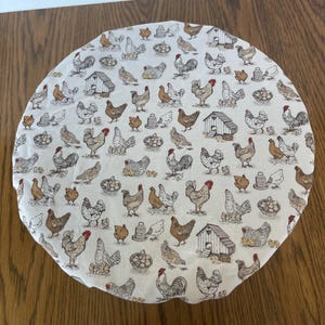 Könnte beinhalten: Ein rundes, weißes Stoffstück mit einem sich wiederholenden Muster aus Hühnern, Hühnerställen und Eiern in Braun-, Grau- und Rottönen. Das Design ist eine charmante Illustration im Landhausstil.