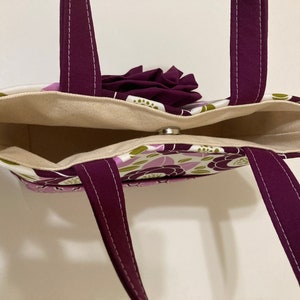 May include: A purple and green floral tote bag with a purple fabric flower on the top. The bag has a cream-colored lining and two purple straps.