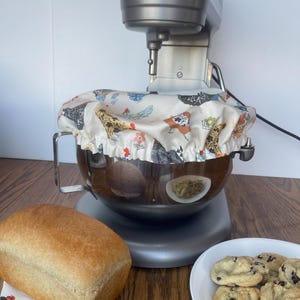 May include: A silver KitchenAid stand mixer with a patterned bowl cover. A loaf of bread sits on a floral coaster, and a plate of chocolate chip cookies is nearby. The mixer is on a wooden surface.