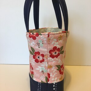 May include: A fabric tote bag with a floral pattern in pink, red, and green. The top of the bag has navy blue handles with white stitching. The bottom of the bag is navy blue with a white dotted design.