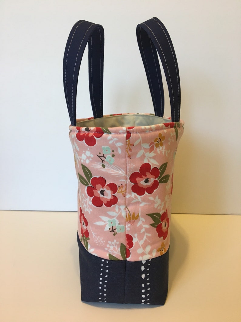 May include: A fabric tote bag with a floral pattern in pink, red, and green. The top of the bag has navy blue handles with white stitching. The bottom of the bag is navy blue with a white dotted design.