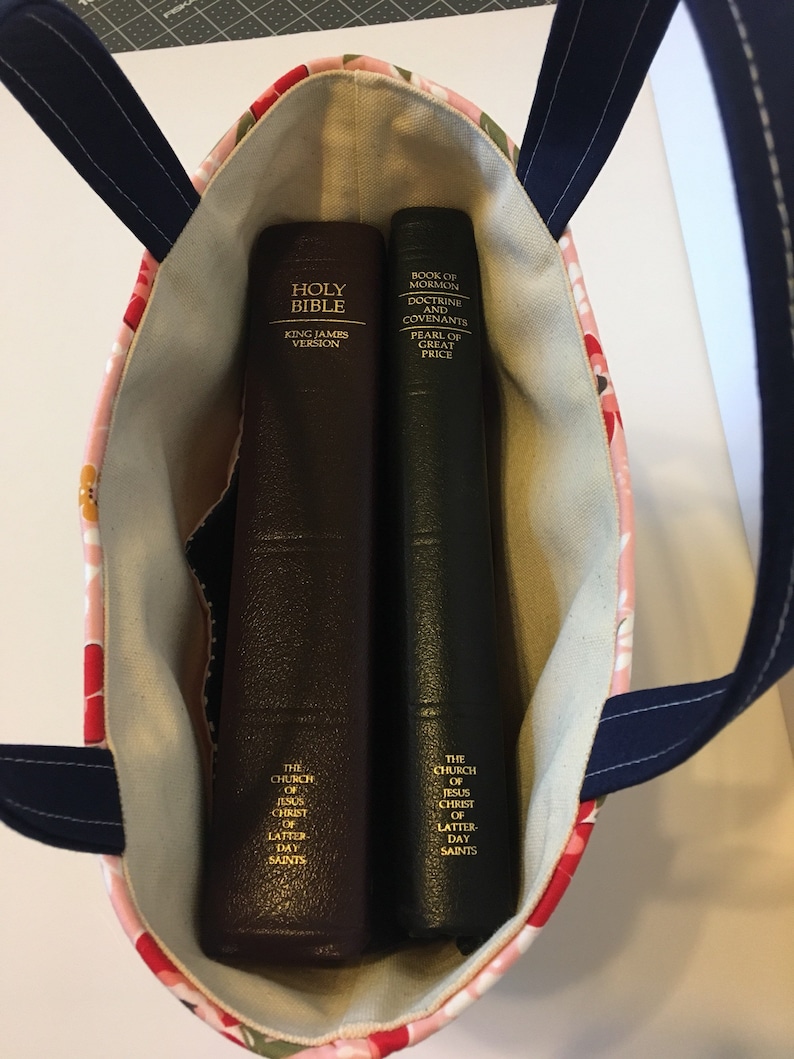 May include: A floral print tote bag with navy blue handles holds two books. One is a brown Holy Bible, King James Version. The other is a dark green Book of Mormon, Doctrine and Covenants, Pearl of Great Price. The bag's interior is a light beige.