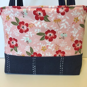 May include: A pink floral tote bag with red flowers, green leaves, and white accents. The bottom section is navy blue with white dotted vertical lines. Navy blue handles complete the design.