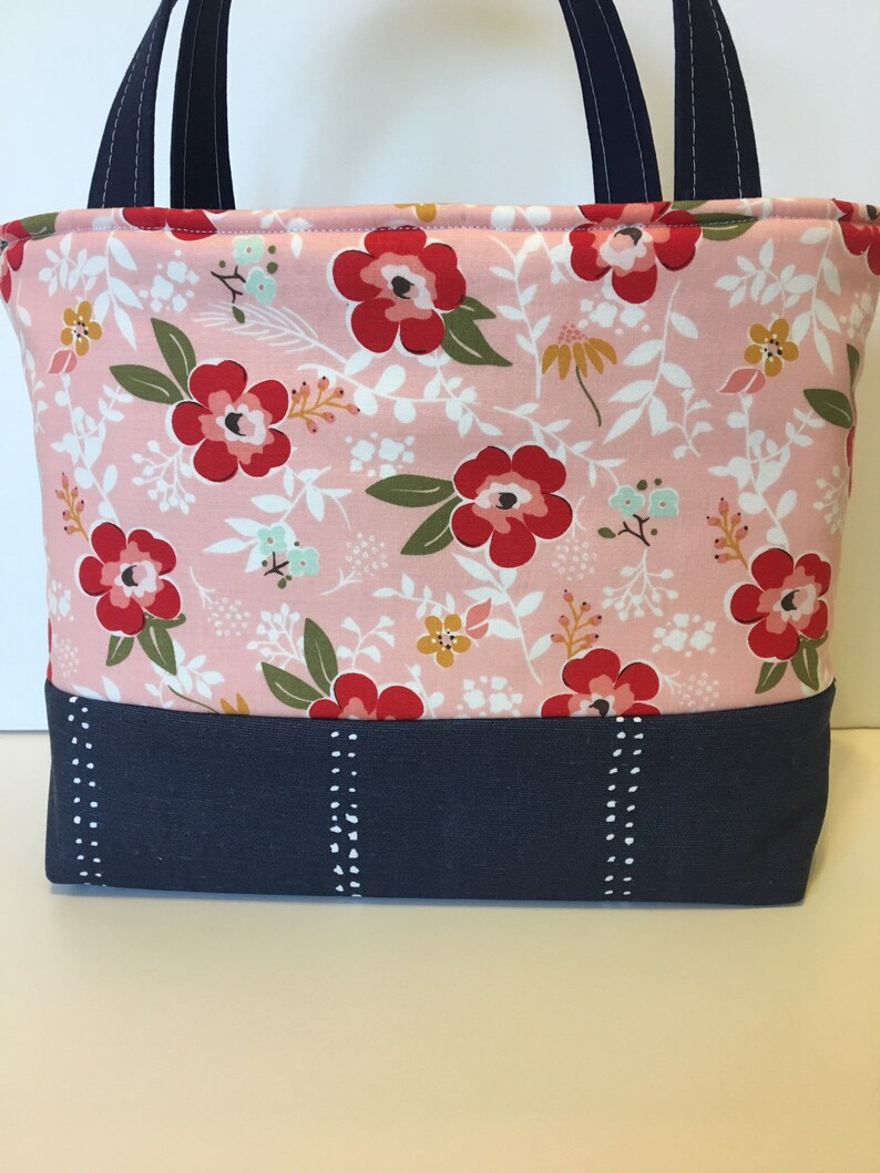 May include: A pink floral tote bag with red flowers, green leaves, and white accents. The bottom section is navy blue with white dotted vertical lines. Navy blue handles complete the design.