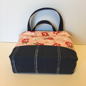 May include: A fabric tote bag with a floral pattern on the top half and a navy blue bottom. The floral pattern features red flowers and green leaves on a light pink background. The bag has navy blue handles and vertical white dotted lines.
