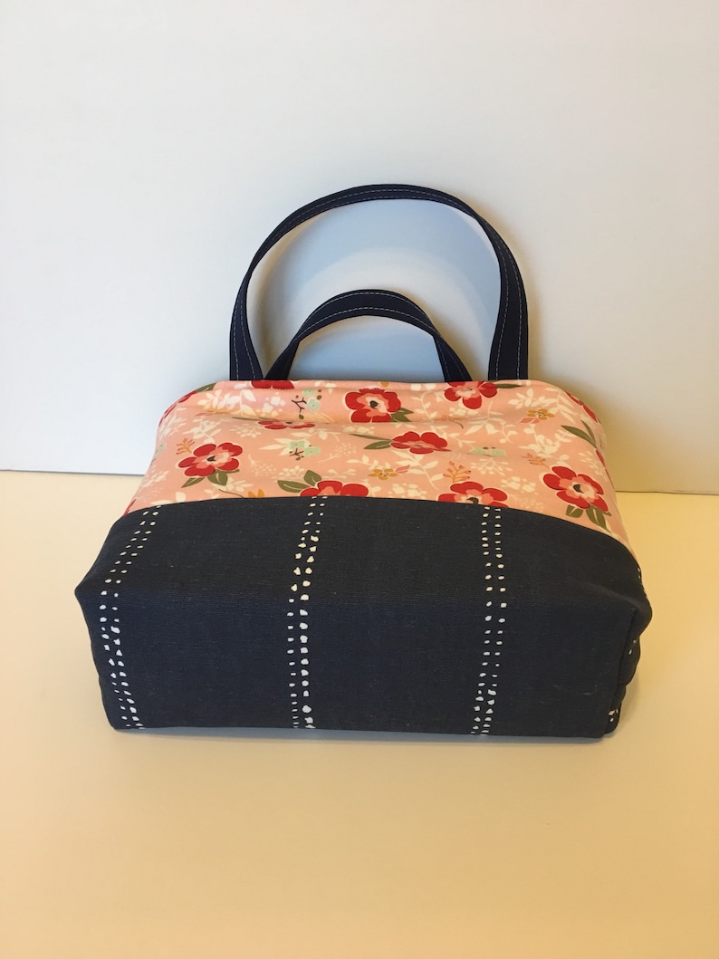 May include: A fabric tote bag with a floral pattern on the top half and a navy blue bottom. The floral pattern features red flowers and green leaves on a light pink background. The bag has navy blue handles and vertical white dotted lines.