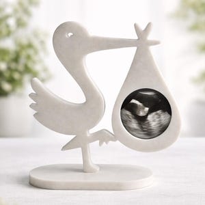 May include: A white, smooth-surfaced stork figurine standing on an oval base. The stork is depicted in profile, holding a teardrop-shaped frame in its beak. Inside the frame is a black and white ultrasound image of a developing fetus.