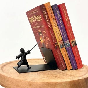 May include: A black metal bookend featuring a sculpted figure in a robe holding a wand pointed towards four Harry Potter books. The books include Harry Potter and the Philosopher's Stone, Chamber of Secrets, Prisoner of Azkaban, and Goblet of Fire.