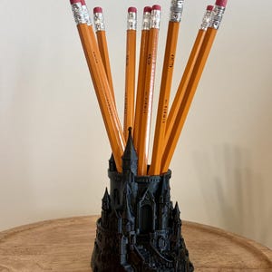 May include: A black 3D-printed pencil holder shaped like a castle with multiple towers and arched doorways. Several yellow HB 2 pencils with pink erasers and silver metal ferrules are inserted into the top of the structure, standing upright.