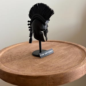 May include: A black miniature Spartan helmet with a tall, textured crest and a decorative tail piece, mounted on a thin black stand. A small rectangular black plaque with the white text THE ODYSSEY is attached to the base of the stand.