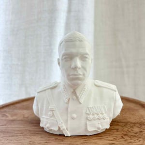 Puede incluir: Un busto blanco impreso en 3D de una persona con un uniforme de estilo militar que incluye charreteras, cuello de camisa, una correa diagonal y varias medallas prendidas en el pecho. La escultura presenta un rostro detallado.