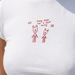 May include: A white short-sleeved t-shirt featuring a red embroidered graphic of two simple cartoon figures and two small clouds. The text "Every cloud has a silver lining" is embroidered in red above the figures in a handwritten style.
