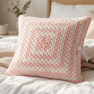 May include: A square throw pillow featuring a crochet granny square pattern in alternating light pink and cream white yarn. The pillow is finished with a small white ribbon bow attached to the side edge, providing a decorative detail to the design.