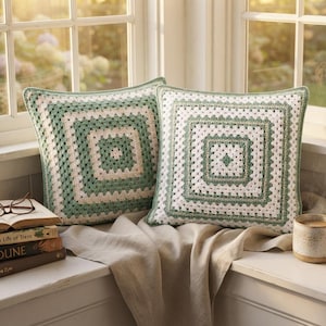 Peut inclure: Deux coussins carrés présentant un motif de granny square au crochet en fil vert sauge et crème alternés. Les coussins sont disposés côte à côte, mettant en valeur un design géométrique classique à l'aspect texturé et artisanal.