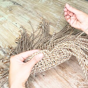 May include: Close-up of a woven basket being crafted. The basket is made of twisted beige and brown rope. The hands are holding the rope and weaving it into the basket. The background is a light-coloured wooden surface.