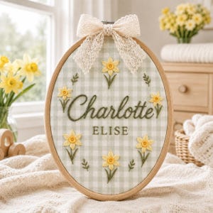 Könnte beinhalten: Ein ovaler Stickrahmen aus Holz mit hellgrün-weiß kariertem Stoff. Der Stoff ist mit gelben Narzissen und den Namen Charlotte und Elise in dunkelgrünem Garn bestickt. Eine cremefarbene Spitzenschleife ist oben am Rahmen befestigt.