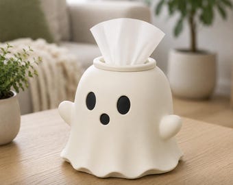 May include: A cream-colored ghost-shaped tissue box cover with two oval black eyes and a small circular black mouth. The top of the ghost features an opening where a white tissue is pulled through, creating a fan-like shape at the top.